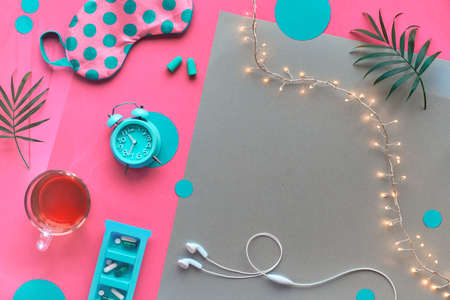 Healthy night sleep creative concept. Sleeping mask, alarm clock, earphones, earplugs, tea and pills. Split pink and craft paper background with light garland and palm leaves. Text space, copy-space.の写真素材
