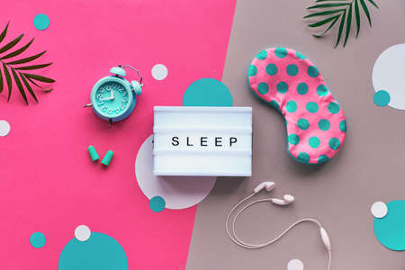 Healthy night sleep creative concept. Sleeping mask, blue mint alarm, earphones, earplugs. Two tone pink and silver background with paper circles and leaves. Flat lay, lightbox with text "Sleep".の写真素材