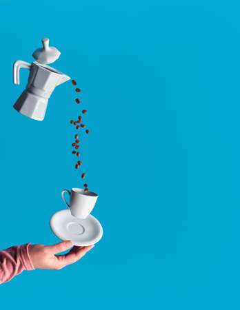 Trendy levitation. Flying line of coffee beans between ceramic coffee maker and espresso cup with saucer held in hand. Blue mint background with copy-space for your text.の写真素材