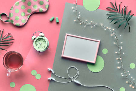 Healthy night sleep creative concept. Sleeping mask, green alarm clock, earphones, earplugs, tea and pills. Split two tone pink silver background with light garland. Copy-space, text space in frame.の写真素材