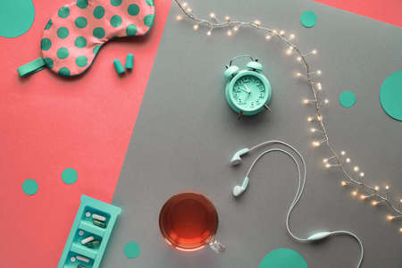 Healthy night sleep creative concept. Sleeping mask, green alarm clock, earphones, earplugs, tea and pills. Split two tone coral color brown background with light garland. Copy-space, text space.の写真素材
