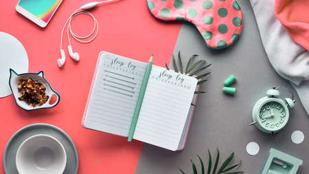 Open sleep log or sleeping diary book. Healthy night sleep creative concept. Sleeping mask, alarm clock, earphones, earplugs, calming tea and pills. Split two tone coral and brown paper background.の写真素材