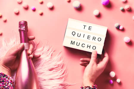 Valentine day creative flat lay, top view. Hands holding lightbox with text and pink bottle of champagne on abstract pink background. Text "Te quiero mucho" means "I love you" in Spanish.の写真素材
