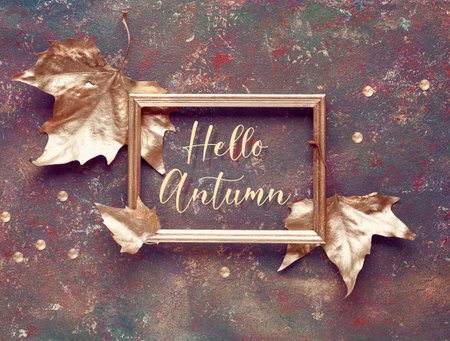 Fall flat lay with sycamore leaves painted gold and gilded text Hello Autumn in golden frame on dark...