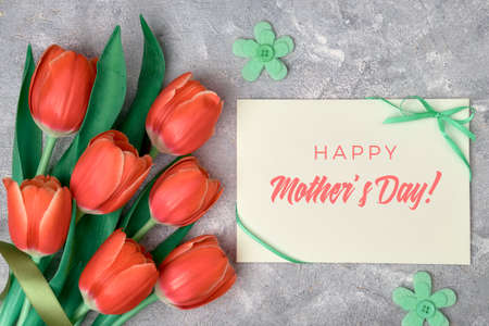 Happy Mother's day, text on paper card with ribbon bow. Flat lay with red tulips, green textile flowers and paper card on grey textured background.の写真素材