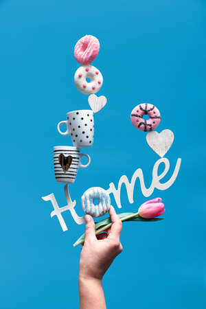 Word "home" with coffee cups, pink and blue donuts and marshmallows. Hand with pink tulip holding pyramid on index finger. Creative Springtime fun life background in blue mint color.の写真素材