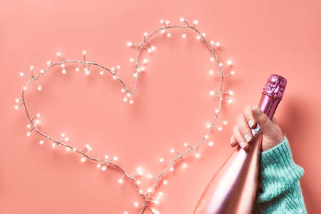 Valentine top view on pink background. Light garland in heart shape, woman hand with metallic pink champagne bottle. St. Valentine's day February 14 concept monochrome flat lay from above.の写真素材