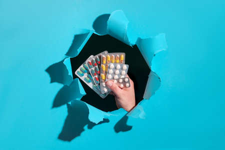 Assorted pills or capsules with medications in hand framed by paper hole. Mint blue trendy torn paper hole background. Selection of medications, food supplements or herbal remedies.の写真素材