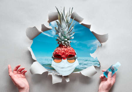Covid-19 coronavirus sudden quarantine in beach resort, cruise ship or hotel. Pineapple wearing sunglasses and medical face mask with sea through ragged paper hole. Hands with disinfectant gel.の写真素材
