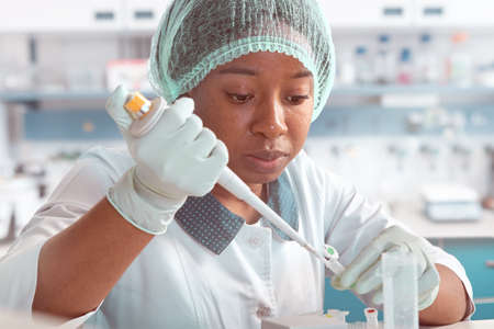 African scientist, technical assistant in lab coat, protective hat and gloves optimizes PCR testing for SARS-CoV-2 virus in modern test laboratory. Troubleshooting of nucleic acid tests.の写真素材