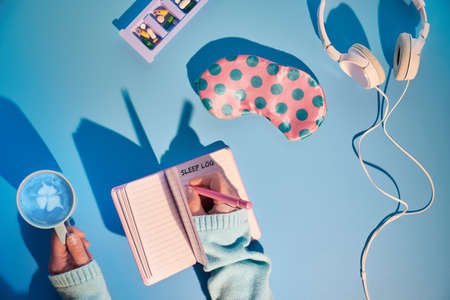Healthy night sleep creative concept in pink and blue. Sleep mask, pink with green polka dots, earphones, calming pills to help anxiety, sleep log notebook. Blue mint background, long shadow design.の写真素材