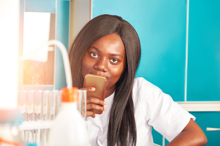 Young smiling African female scientist, graduate or medical student with mobile phone looks at the...