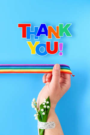 Rainbow ribbon in woman's hand. Chamomile flowers and grass bouquet attached to wrist with medical patch. Thank you doctors and nurses. Creative flat lay, top view on blue background.の写真素材