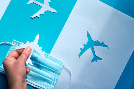 Hand holding paper plane over face mask takes it from shadow to light. Air travel resumes after...