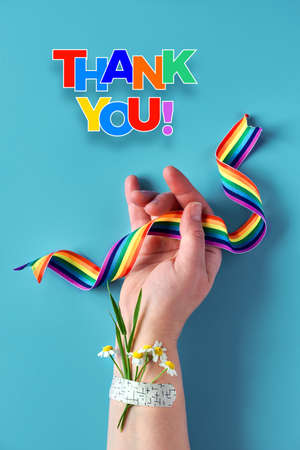 Thank you doctors and nurses! Rainbow ribbon in hand of mature woman with chamomile flower and grass bouquet attached with medical aid patch. Creative flat lay, top view on blue background.の写真素材