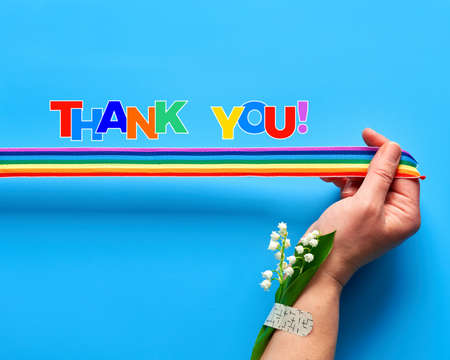 Thank you doctors and nurses. Rainbow ribbon in woman's hand. Chamomile flowers and grass bouquet attached to wrist with medical patch. Creative flat lay, top view on blue background.の写真素材