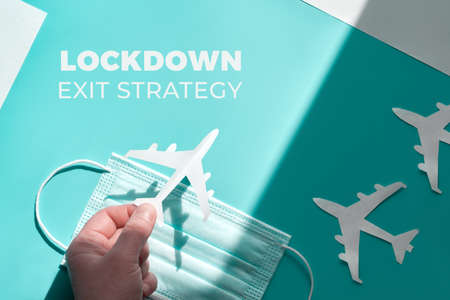 Hand holding paper plane over face mask from shadow to light. Air travel resumes after travel. Vacations stopped and borders closed during coronavirus pandemics. Text Lockdown exit strategy.の写真素材
