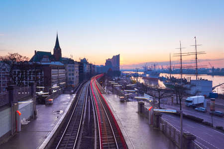 Landungsbrucken station in Hamburg, Germany on a sunrise, early in the morning, with tracer of rear lights of passing train. Elbe riverside, downtown skyline on sunrise.のeditorial素材