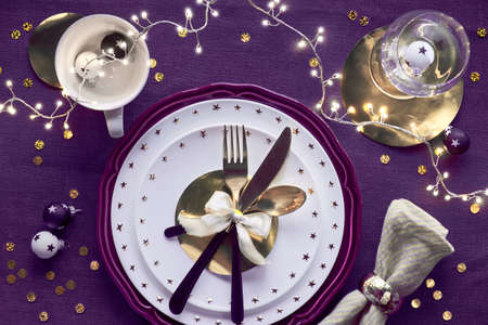 Christmas table setup with white plate and golden utensils and gilded decorations on deep violet table cloth. Flat lay, top view on dark purple linen textile background. Xmas lights garland.の写真素材