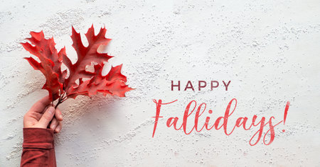 Hand holding twig with vibrant red oak leaves, text Happy Fallidays. Panoramic flat lay on white...