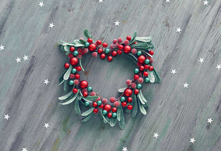 Decorative Xmas mistletoe wreath with red berries and green leaves in shape of heart. Wooden Christmas background decorated with paper star confetti.の写真素材