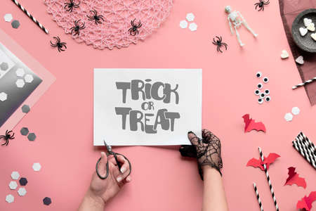 Trick or treat text on pink paper background. Flat lay, hands with scissors and Halloween decor. Top view on hexagon confetti, paper drink straws, bats, sugar hearts and spiders.の写真素材
