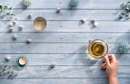 Wintertime tea, flat lay arrangement with glass teapot, glass of tea in hand on faded blue wooden planks. Christmas decorations on the table - mirror balls, baubles, toys, tea candle and eucalyptus.の写真素材
