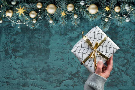 Christmas flat lay on dark green wood. Hand holding wrapped gift. Fir twigs with gold decor.の写真素材