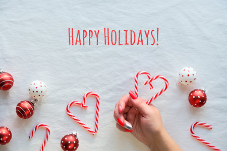 Christmas background, flat lay, top view on white textile background. Female hands holding heart shapes made from candy canes. Red and white trinkets with dots.の写真素材