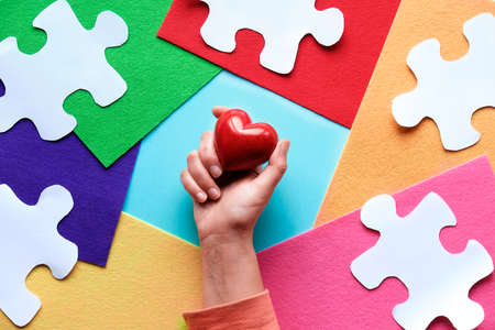 Hands holding red stone heart. Creative design for April 2, Autism World Awareness day. Jigsaw puzzle element on layered colorful felt background. Flat lay, top view, creative concept.の写真素材