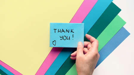 International Thank you day. Greeting card with female hand holding envelope with heart shape and flowers. Thank you handwritten card in hand. Creative flat lay colored layered paper background.の写真素材
