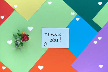 International Thank you day. Greeting card with female hand holding envelope with heart shape and flowers. Thank you handwriting on card. Concept flat lay on multicolor layered paper background.の写真素材