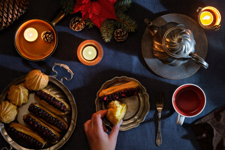 Yummy eclairs or brewing cakes and pewter tea pot and cup of red tea. Natural Christmas decor, candles, red poinsettia on fir twigs. Xmas top view, flat lay on dark purple linen textile tablecloth.の写真素材