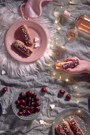 Happy Valentine celebration Creative flat lay with assortment of yummy eclairs or brewing cakes and cherry on textile background with pink fur mat and light garland. Hand holding half eaten eclair.の写真素材