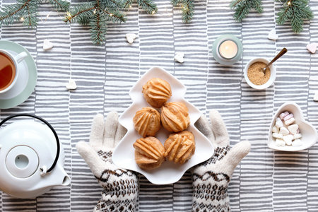 Creative Xmas table setup for tea. Top view with eclairs, brewing cakes. Hands in gloves show plate with cakes in shape of Christmas tree. Fir twigs with light garland on stripy textile tablecloth.の写真素材