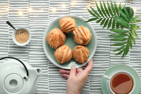 Creative flat lay with yummy eclairs or brewing cakes. White ceramic tea pot, cup of tee, sugar. Hand holds plate with cakes. Palm leaves in vase, light garland on light stripy textile tablecloth.の写真素材
