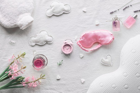 Healthy night sleep creative concept in pink and green. Off white, ivory textile background. Pink hyacinth flowers, alarm clock. Orthopedic pillow, sleep mask, tea, candle, warm bottle.の写真素材