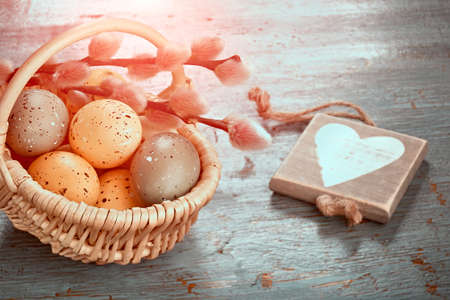 Easter decorations, toned image. Wattle rattan basket with two tone Easter eggs, green and orange. Close-up on pussy willow twigs. Wooden tag with heart shape. Toned greeting decorations with flare.の写真素材
