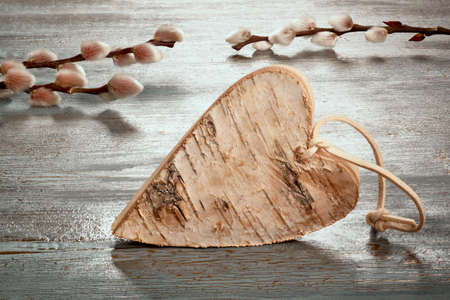 Easter decorations, toned image in desaturated earth colors. Close-up on rustic heart made from birch wood. Pussy willow twigs. Rustic wooden table with textured painted surface in counterligt.の写真素材