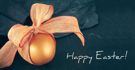 Single golden Easter egg with orange bow on dark background. Happy Easter text on black slate stone. SImple panoramic Easter greeting card design.の写真素材