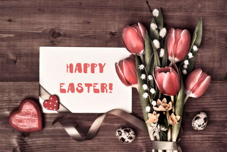 Card with text Overhead view, flat lay. Arrangement of Easter, springtime decorations. Wrapped gifts, red tulips, pussy willow, lily of valley flowers. Easter eggs, hearts, ribbons on brown wood.の写真素材
