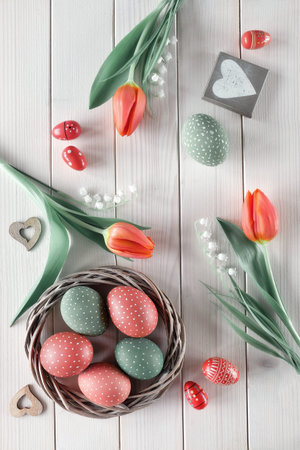 Flat lay with red tulips in rattan wreath basket and green grass. Decorative painted Easter eggs....