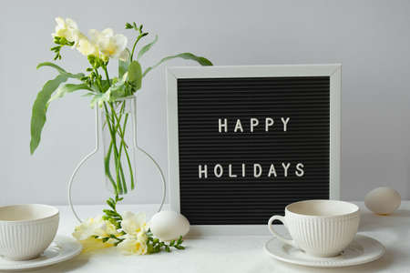 Text Happy Holidays on letter board with spring freesia flowers and tea cup on table. Black letterboard with text. Natural Easter eggs, white ceramic cups and flowers on grey background.の写真素材