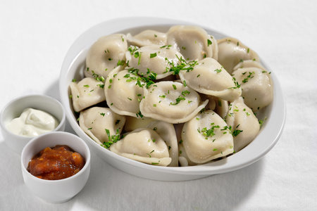 Boiled or steamed pelmeni in white bowl. Pelmeni are homemade pasta with mince meet filling wrapped in thin wheat dough. Ajika and sour cream sauce in small ceramic pinch bowls on the side.の写真素材