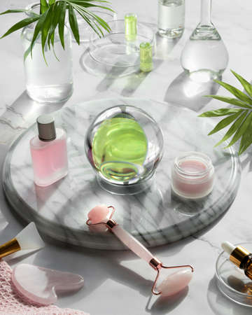 Gua Sha massager and pink quarz face roller on marble stone background. Face care products, glass crystal ball and exotic palm leaves. Towel, skincare tonic, essential oil and revitalizing serum.の写真素材