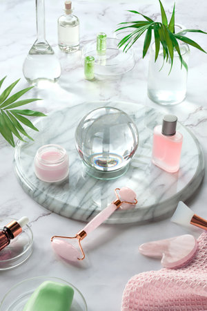 Gua Sha massager and pink quarz face roller on marble stone background. Face care products, glass crystal ball and exotic palm leaves. Towel, skincare tonic, essential oil and revitalizing serum.の写真素材