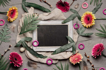 Jungle floral flat lay, linen textile. Blackboard, chalk board with copy-space. Tropical plants, with burgundy, orange, white gerbera flowers, feathers, quail eggs. Flat lay, exotic fern leaves.の写真素材
