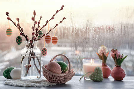 Easter decorations on windowsill in Spring. Wooden painted eggs on pussy willow twigs, Easter eggs, hyacinth bulbs in wax, macrame mats. Window with urban view, gray rainy day, rain drops on window.の写真素材