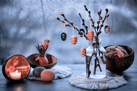 Evening window in Spring, wooden Easter eggs on pussy willow twigs in glass bottle, window with blue evening light and rain drops, candle and painter Easter egg with ribbon.の写真素材