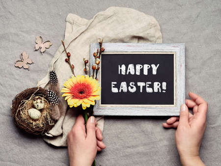 Happy Easter, chalk text on black board. Hands holding blackboard and orange gerbera flower. Quail eggs in bird nest, spring decor. Flat lay, linen textile background. Easter greeting card designの写真素材